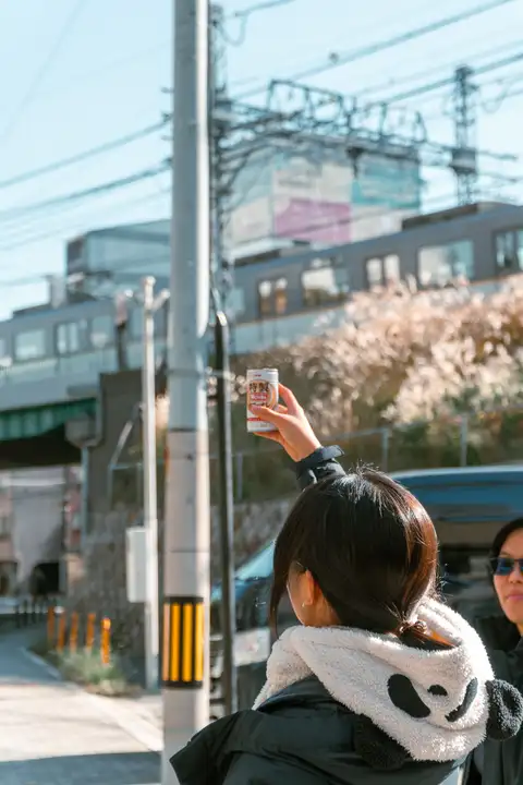 The photographer's sister holding up a canned coffee beneath cherry blossoms on a sunny street