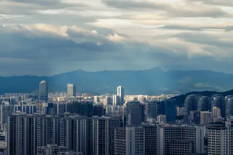 Late sunlight crossing a dense city beneath dark mountains and layered clouds
