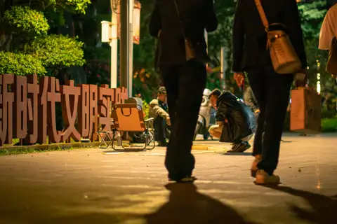 Pedestrians crossing warm streetlight while people gather beside folding chairs