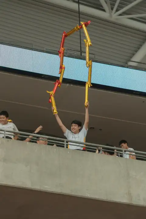 A smiling student raising linked red and gold inflatable sticks above a stadium railing
